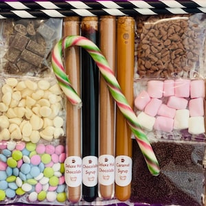 Puede incluir: Una caja de regalo llena de ingredientes para chocolate caliente. Incluye chispas de chocolate, malvaviscos, chispas, chocolates recubiertos de caramelo, chispas de chocolate blanco y tubos de jarabe de chocolate y caramelo. Un bastón de caramelo está en el centro.