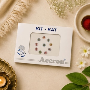 May include: A white rectangular product packaging labelled KIT-KAT and Accron, featuring a central window displaying nine small, circular, multi-coloured decorative bindi stickers arranged in a circular pattern with a blue rose illustration on the left.