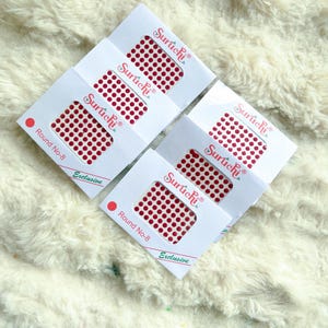 May include: Four white card packs of red dot stickers, labelled "Suruchi" and "Round No-8 Exclusive". The stickers are arranged in a grid pattern. The background is a fluffy, off-white surface.