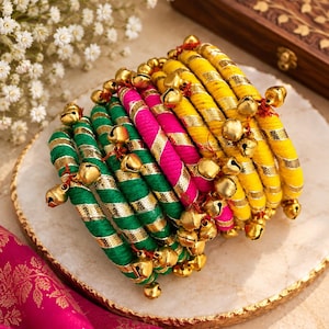 May include: A set of stacked bangles wrapped in vibrant green, hot pink, and yellow thread, each accented with a spiralling metallic gold ribbon and small, dangling gold-toned bells attached with thin red thread throughout the circular jewellery.