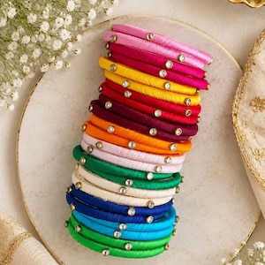 May include: A stack of multicolored bangles wrapped in silk thread, each featuring small, clear, round embedded stones. The collection includes shades of pink, yellow, red, orange, light pink, green, white, dark blue, and bright blue.
