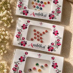 May include: Three rectangular white cardboard packages of Ambika Fancy Bindi stickers. Each package features a floral border and a central window displaying small, colourful, circular adhesive bindis in various shades and designs.