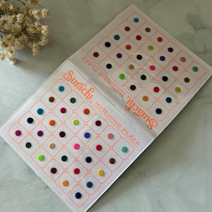 May include: A sheet of colourful, round, adhesive bindi stickers. The sheet is divided into a grid of 49 squares, each containing a single bindi. The bindis are in a variety of colours, including red, green, blue, pink, and yellow. The sheet is labelled "Surachu Matching Plaza".