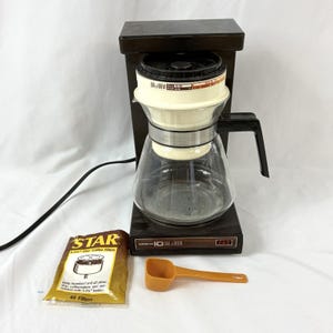 May include: A vintage black and cream-colored coffee maker with a glass carafe. Includes a package of coffee filters and an orange scoop. The coffee maker has the text "Norelco Dial a Brew".