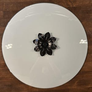 May include: A circular, clear glass lid featuring a central black metal handle shaped like a stylized flower with multiple petals. The lid has a smooth, reflective surface and a thin, dark rim, designed to fit securely over a corresponding container.