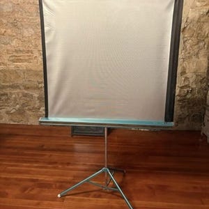 May include: A portable projection screen featuring a rectangular white fabric surface with black borders, mounted on a teal metal housing. The screen is supported by a collapsible silver metal tripod stand with three legs and white rubber feet.