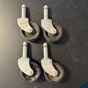 May include: A set of four swivel casters featuring black rubber wheels and silver-toned metal mounting brackets with vertical stems. Each caster shows signs of oxidation and wear on the metal surfaces, with visible screws securing the wheels.