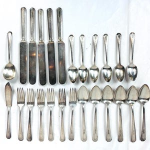 May include: A collection of silver-toned flatware including five dinner knives, one butter knife, six forks, and twelve spoons of varying sizes. The utensils feature simple, polished handles and metallic surfaces, arranged in rows on a plain white background.