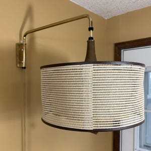 May include: A mid-century modern wall-mounted lamp with a brass arm and a cylindrical lampshade. The lampshade is made of a woven, off-white material with dark brown trim. The lamp is mounted on a beige wall.