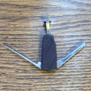 May include: A multi-tool featuring a dark brown wooden handle with three metallic implements extended outward. The central component is a small metal pick, while the two side pieces are thin, flat metal blades with pointed tips.