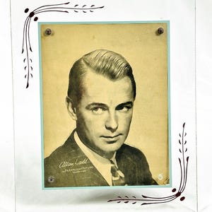 May include: A vintage glass picture frame with a black and white portrait of Allan Ladd. The frame has a light blue border and decorative brown floral accents. The text "Allan Ladd in Paramount Pictures" is visible.