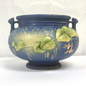 May include: A matte blue ceramic bowl with two side handles and a pedestal base. The exterior features a raised relief design of light green leaves and pale yellow floral elements against a textured blue background with visible wear on the rim.