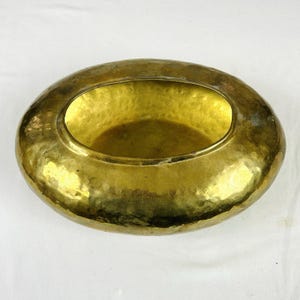 May include: An oval-shaped brass bowl featuring a distinct hammered texture across its entire exterior and interior surface. The metal has a polished, reflective gold-toned finish with subtle variations in color and light throughout the piece.