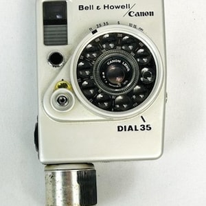 May include: A silver Bell & Howell/Canon Dial 35 camera featuring a unique circular lens array and a prominent silver film advance knob at the base. The front body displays the brand names and the model name DIAL 35 in black text.