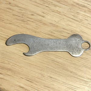 May include: A vintage, silver-colored bottle opener with a seal-like design on one end and a small ring on the other. The opener has a curved shape with a star and lines etched into the seal's head. The tool is made of metal and shows signs of wear.
