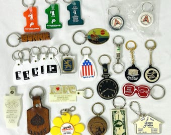May include: A collection of various promotional keychains featuring diverse shapes, materials, and designs. Items include plastic number ones, a wooden Bonnie tag, a clock face, a flower, a house shape, and several circular and rectangular metal or plastic tokens.