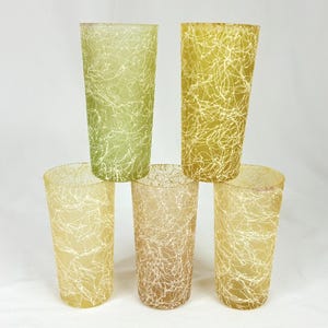 May include: A set of five cylindrical drinking glasses featuring a textured, frosted finish in varying shades of green and yellow. Each glass is decorated with a white, chaotic, string-like pattern that wraps around the exterior surface.