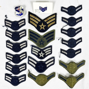May include: A collection of military rank insignia patches and one metal pin. The patches feature chevron designs in various colors including dark blue, olive green, and tan with embroidered stars and stripes. One small white card displays a pin.