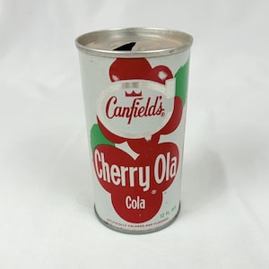 May include: Vintage Canfield's Cherry Ola Cola can. The can is cylindrical with a silver top and bottom. The label features a red cherry design with the words "Canfield's Cherry Ola Cola" in red and white. The can is 12 fl oz.