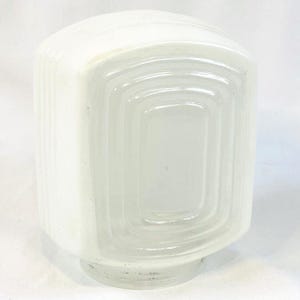 May include: A vintage, opaque white glass light fixture shade. The shade has a rectangular shape with rounded corners and a series of concentric, recessed rectangles on the front. The sides have vertical ridges. The base is a small, circular foot.
