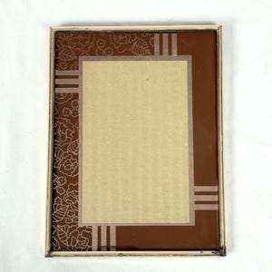 May include: Rectangular picture frame with a brown border and a beige interior. The frame has a floral pattern on the left side and vertical stripes on the top and bottom. The frame is made of wood.