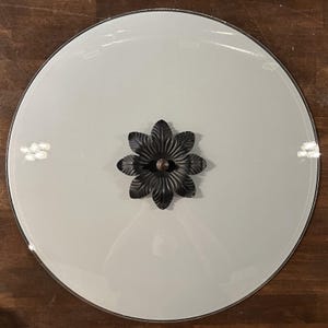 May include: A circular glass mirror with a smooth, reflective surface and a beveled edge. Centered on the mirror is a decorative black metal accent piece shaped like a six-petaled flower with textured, ribbed leaves and a small central knob.