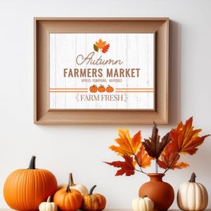 Pode incluir: Uma impressão emoldurada com um fundo de madeira branca e o texto "Autumn Farmers Market" em letras marrons. O texto "Apples Pumpkins Hayrides" está abaixo do texto principal em letras marrons menores. O texto "Farm Fresh" está abaixo do texto menor em letras marrons com um design de folha estilizado de cada lado. Há três abóboras laranja com caules verdes em fila abaixo do texto. A impressão é cercada por uma moldura marrom.