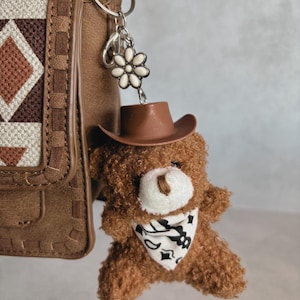 May include: A brown teddy bear keychain wearing a cowboy hat and a bandana with a black and white pattern. The keychain is attached to a brown purse with a geometric design.