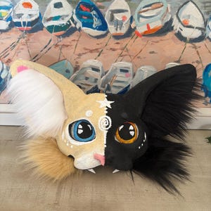 May include: A split-faced cat mask with a cream and black design. The cream side has a pink nose, blue eyes, and a white ear with pink accents. The black side has orange eyes and a black ear. The mask has faux fur.