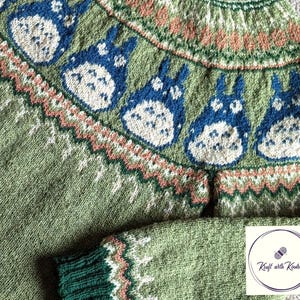 May include: A green knitted sweater featuring blue and white cartoon characters across the yoke. The sweater has a green body, with a green and white patterned cuff. The yoke has a pattern of peach, white, and green. The sweater is handmade.