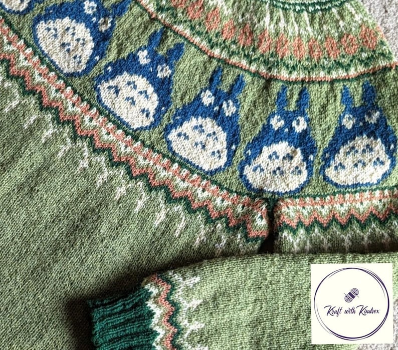 May include: A green knitted sweater featuring blue and white cartoon characters across the yoke. The sweater has a green body, with a green and white patterned cuff. The yoke has a pattern of peach, white, and green. The sweater is handmade.
