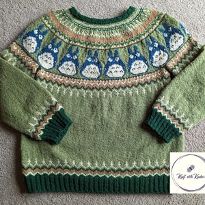 May include: A green knitted sweater featuring a pattern of blue and white cartoon characters. The sweater has a round neck, long sleeves, and ribbed cuffs and hem in a darker green. The design includes geometric and floral patterns in shades of green, pink, and white.