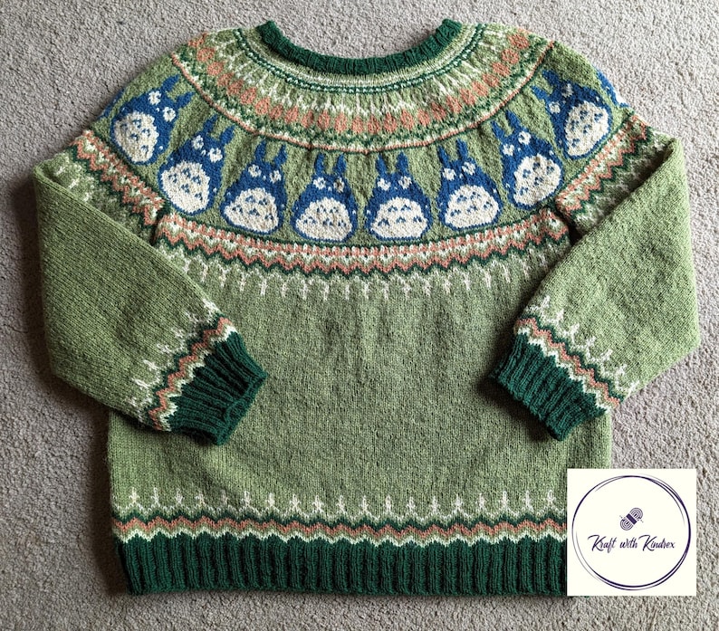 May include: A green knitted sweater featuring a pattern of blue and white cartoon characters. The sweater has a round neck, long sleeves, and ribbed cuffs and hem in a darker green. The design includes geometric and floral patterns in shades of green, pink, and white.