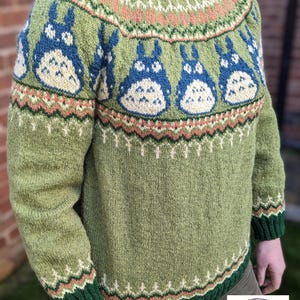 May include: Green knit sweater featuring blue and cream cartoon characters. The sweater has a green collar and cuffs, with patterned details in peach, cream, and green. The design includes a repeating pattern of the cartoon characters across the chest.