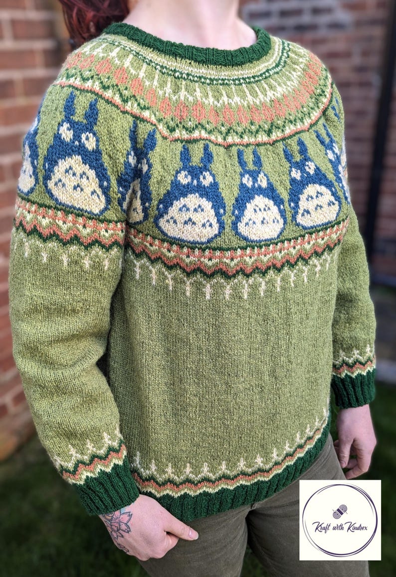May include: Green knit sweater featuring blue and cream cartoon characters. The sweater has a green collar and cuffs, with patterned details in peach, cream, and green. The design includes a repeating pattern of the cartoon characters across the chest.
