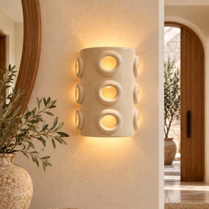 Op de afbeelding: Een halfcilindervormige wandlamp van gebroken wit keramiek, voorzien van drie verticale rijen cirkelvormige uitsparingen die een warm, gloeiend licht uitstralen. Het armatuur is vlak tegen een gestructureerde muur gemonteerd en verspreidt zacht licht.