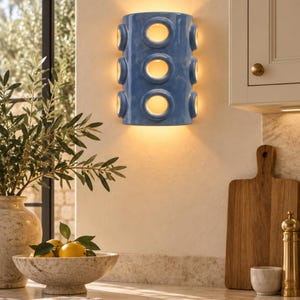 Op de afbeelding: Een cilindervormige wandlamp met een glanzende blauwe afwerking, voorzien van drie verticaal geplaatste ronde uitsparingen aan de voorzijde en extra ronde openingen aan de zijkanten, die een warme gloed van binnenuit verspreidt.
