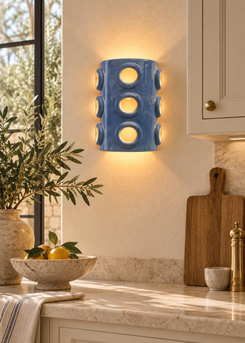 Op de afbeelding: Een cilindervormige wandlamp met een glanzende blauwe afwerking, voorzien van drie verticaal geplaatste ronde uitsparingen aan de voorzijde en extra ronde openingen aan de zijkanten, die een warme gloed van binnenuit verspreidt.