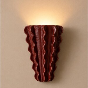 Puede incluir: Un aplique de pared de cer&aacute;mica de color rojo oscuro con un dise&ntilde;o vertical ondulado y plisado. El objeto tiene un acabado brillante y emite un brillo c&aacute;lido y suave desde la abertura superior, proyectando luz hacia arriba.