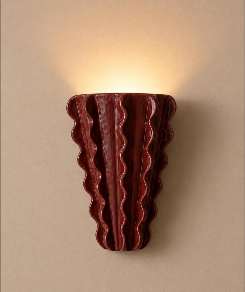 Puede incluir: Un aplique de pared de cer&aacute;mica de color rojo oscuro con un dise&ntilde;o vertical ondulado y plisado. El objeto tiene un acabado brillante y emite un brillo c&aacute;lido y suave desde la abertura superior, proyectando luz hacia arriba.