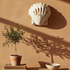 May include: A white ceramic wall sconce shaped like a seashell with vertical cutouts, mounted on a solid wall. The fixture emits a warm light through its openings, casting a distinct shadow on the textured surface behind it.