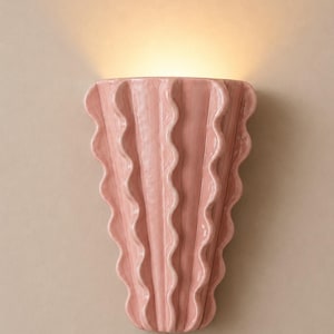 Puede incluir: Un aplique de pared con un acabado de cerámica rosa claro brillante. La lámpara presenta una forma vertical en abanico con crestas onduladas y festoneadas repetitivas en su superficie, emitiendo un suave resplandor cálido desde la parte superior.