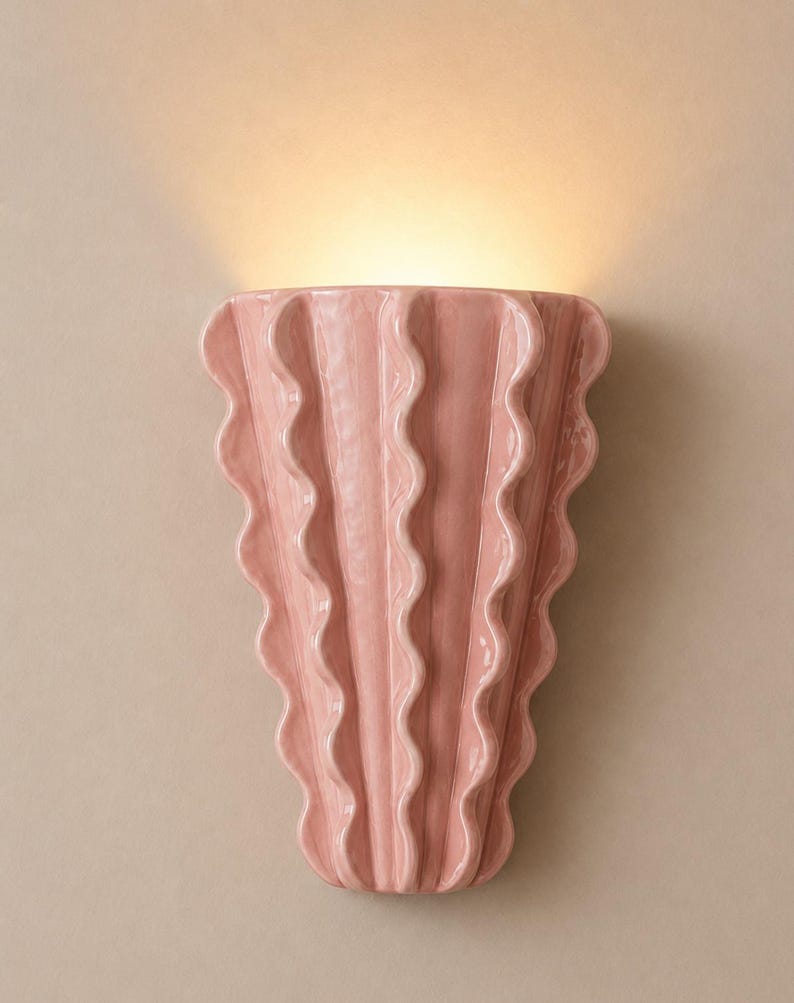 Puede incluir: Un aplique de pared con un acabado de cer&aacute;mica rosa claro brillante. La l&aacute;mpara presenta una forma vertical en abanico con prominentes crestas onduladas que se repiten a lo largo de su superficie, emitiendo una luz c&aacute;lida desde la parte superior.