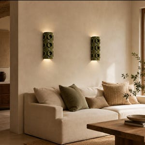 Op de afbeelding: Twee cilindrische olijfgroene keramische wandlampen met een herhalend cirkelvormig uitgesneden patroon, gemonteerd op een beige muur. Elke lamp verspreidt een warme gloed naar boven en beneden, wat de muur boven een cr&egrave;mekleurige bank verlicht.