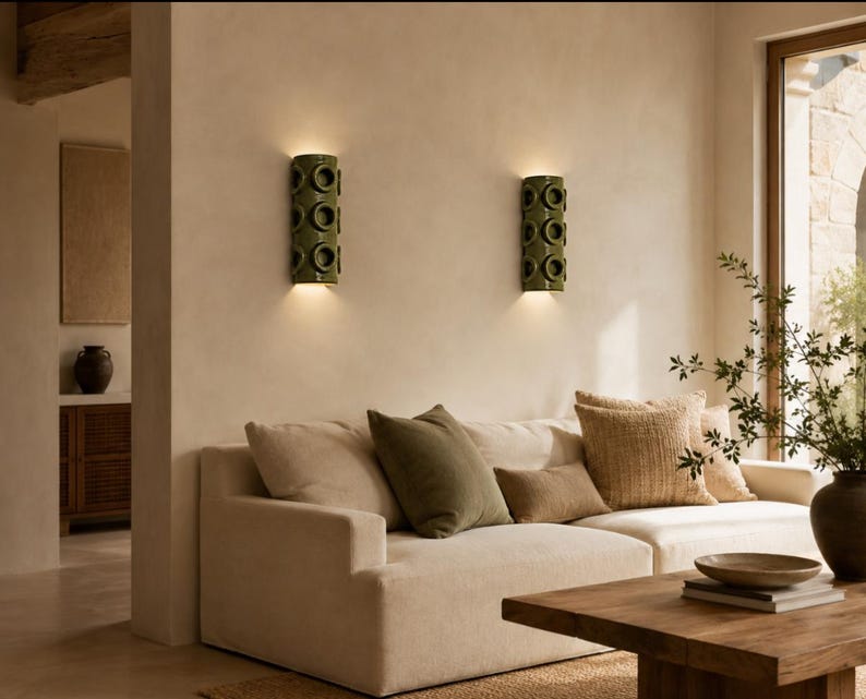 Op de afbeelding: Twee cilindrische olijfgroene keramische wandlampen met een herhalend cirkelvormig uitgesneden patroon, gemonteerd op een beige muur. Elke lamp verspreidt een warme gloed naar boven en beneden, wat de muur boven een cr&egrave;mekleurige bank verlicht.