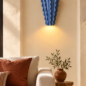 Puede incluir: Un aplique de pared de cer&aacute;mica azul con un dise&ntilde;o vertical ondulado en forma de abanico. La l&aacute;mpara est&aacute; montada en la pared y emite una luz c&aacute;lida y brillante tanto por el borde superior como por el inferior, proyectando una luz suave.