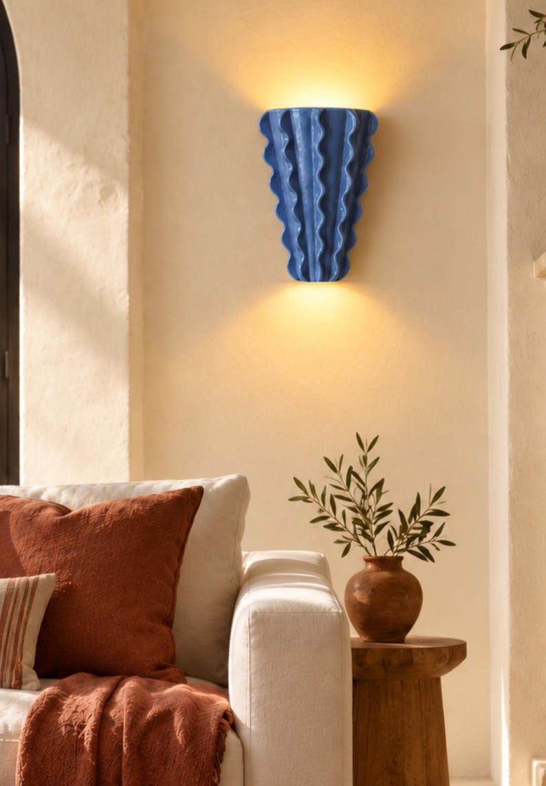 Puede incluir: Un aplique de pared de cer&aacute;mica azul con un dise&ntilde;o vertical ondulado en forma de abanico. La l&aacute;mpara est&aacute; montada en la pared y emite una luz c&aacute;lida y brillante tanto por el borde superior como por el inferior, proyectando una luz suave.