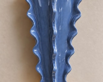 Puede incluir: Un aplique de pared de cerámica azul con una cresta central vertical y bordes ondulados y fruncidos. La lámpara emite un brillo cálido y suave desde las aberturas superior e inferior, iluminando la pared tras la estructura curva.