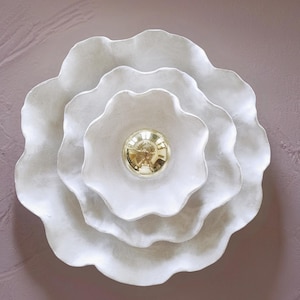 Könnte beinhalten: Weiße, mehrschichtige, blütenförmige Wanddekoration. Die Blütenblätter haben einen strukturierten, wellenförmigen Rand. Die Mitte hat einen goldfarbenen, kugelförmigen Akzent. Die Dekoration ist an einer gedämpften, mauvefarbenen Wand angebracht.