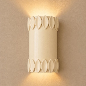 May include: A cylindrical wall sconce in a cream-coloured finish, featuring a smooth central body flanked by decorative leaf-shaped accents at the top and bottom edges. The fixture emits a warm, soft glow from both the top and bottom openings.